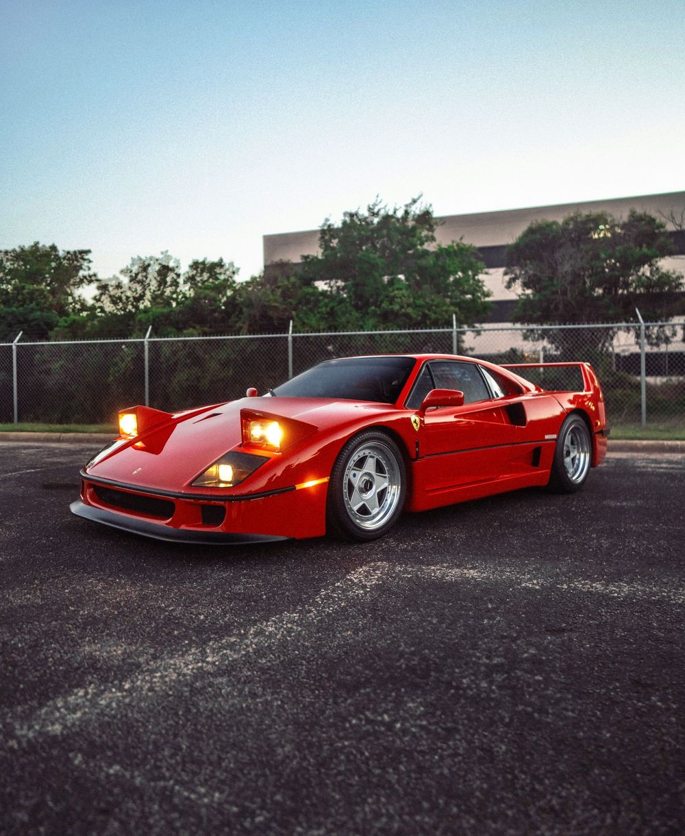 Red Ferrari F40 supercar parked in a lot at dusk, with headlights on and a dramatic low-angle perspective.