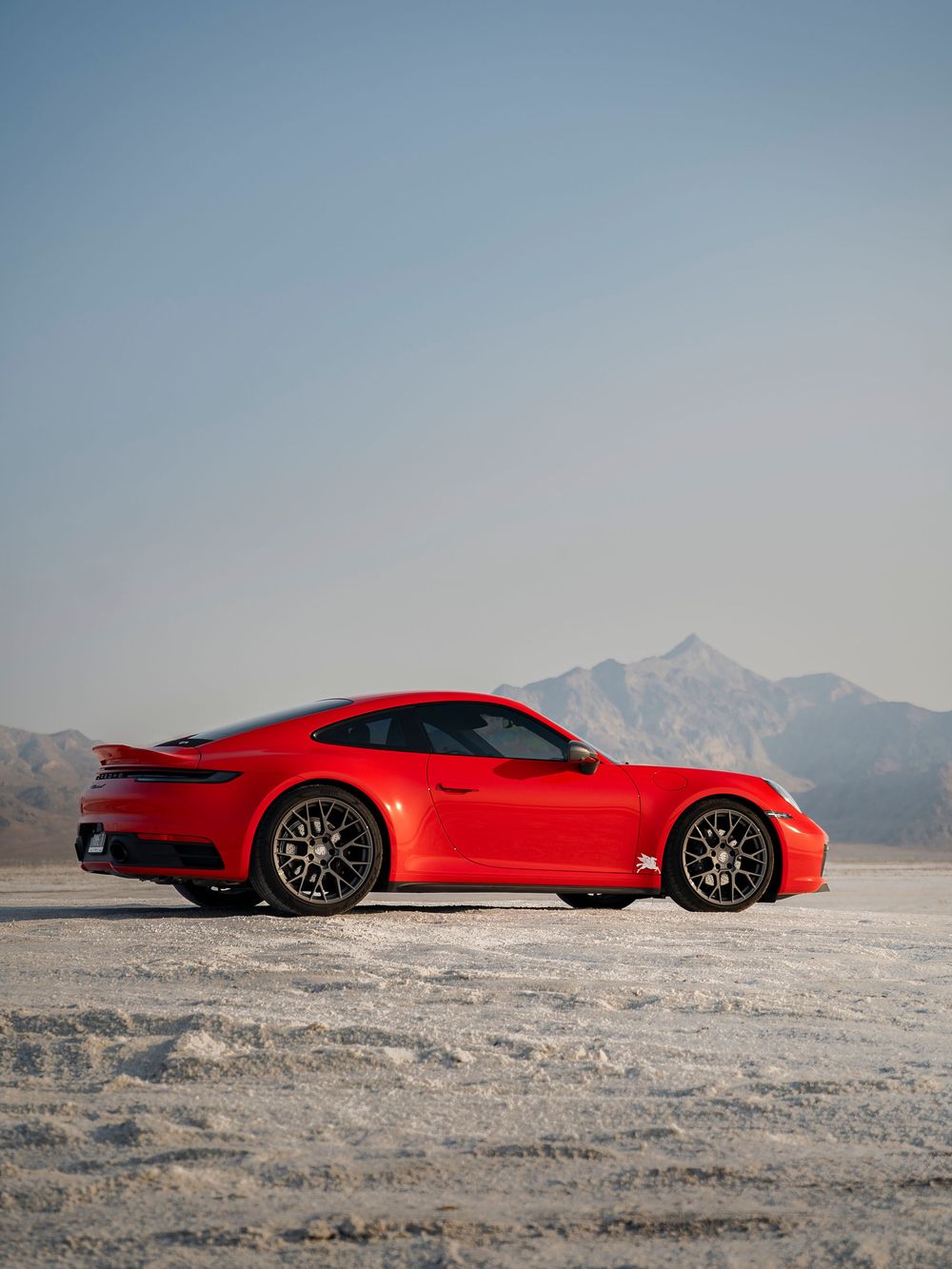 Red Porsche 911 Carrera parked on salt flats with mountain backdrop under clear blue sky.