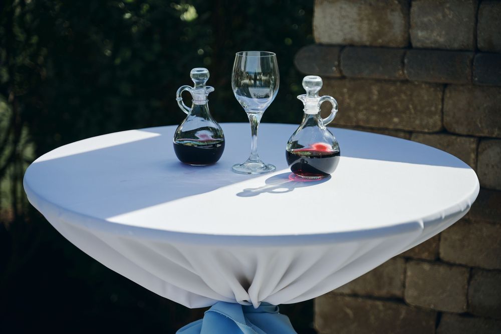 Elegant cocktail table with white cloth, wine glass, and decorative glass decanters set outdoors for a wedding ceremony.