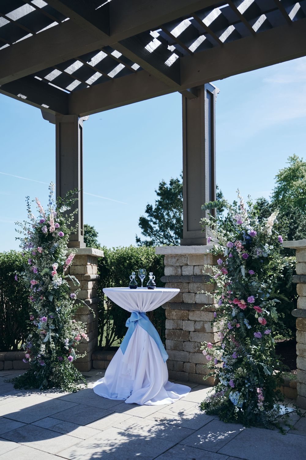 Elegant outdoor wedding ceremony setup with floral arrangements, draped cocktail table, and pergola against a blue sky.