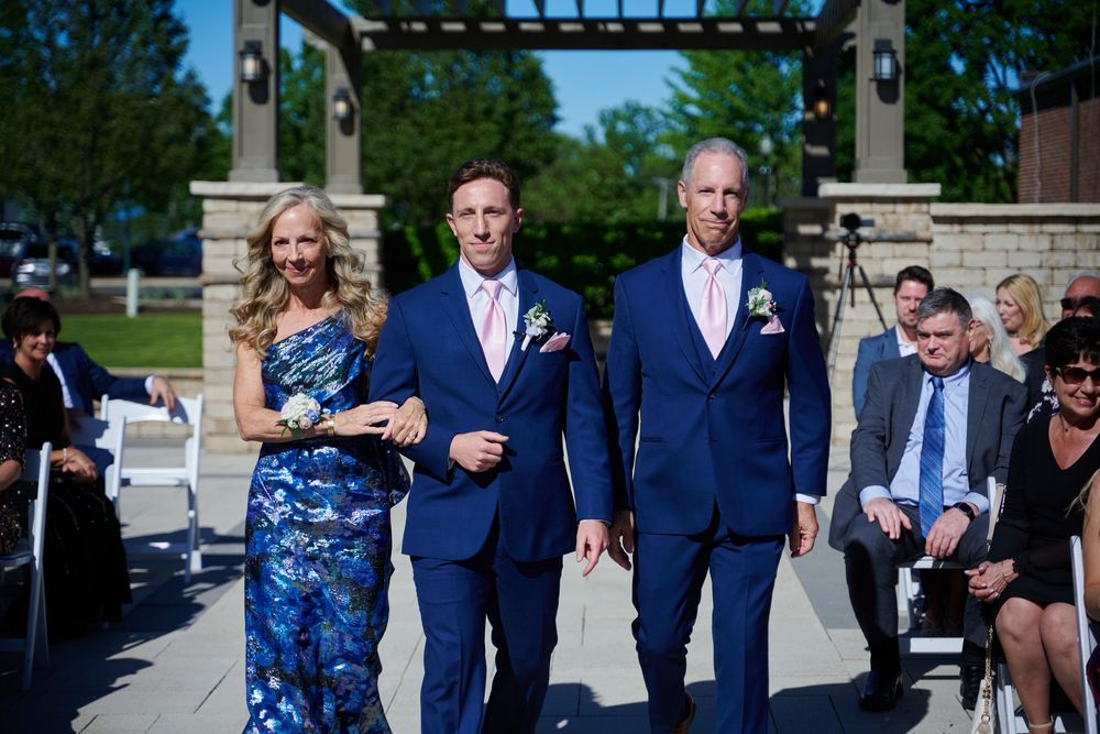 Groom walks down outdoor wedding aisle escorted by parents in navy suits and floral dress on sunny day.