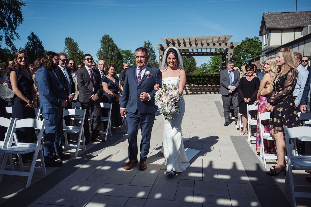 Bride walks down outdoor wedding aisle with escort, guests seated in white chairs, pergola backdrop on sunny day.
