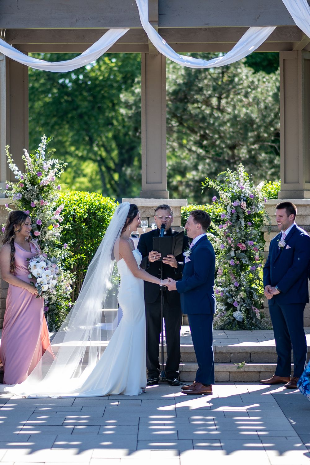 Bride and groom exchange vows at outdoor wedding ceremony with floral arch, officiant, bridesmaid, and groomsman under pavilion.