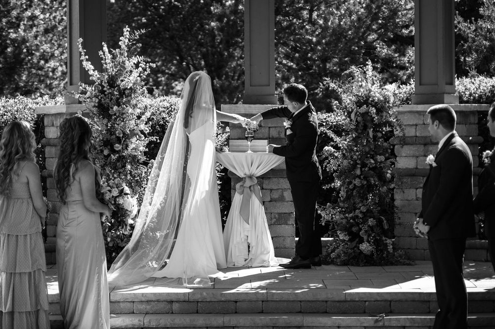 Black and white outdoor wedding ceremony with bride in long veil, groom, officiant, and bridesmaids at stone pavilion.