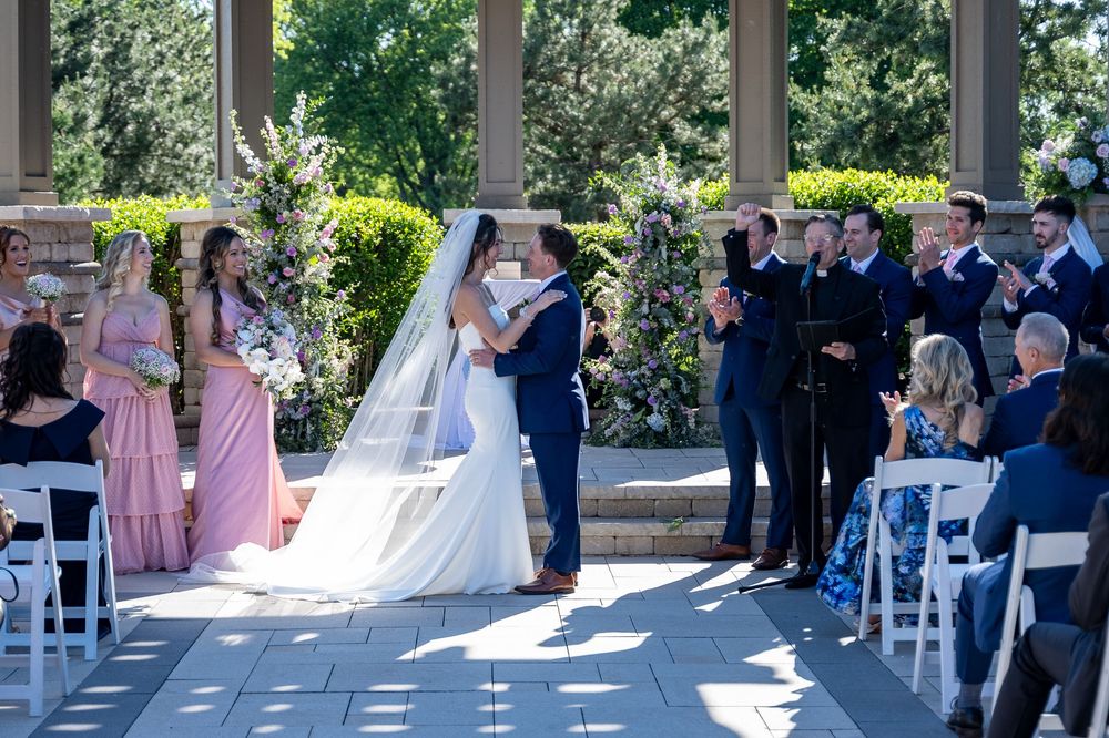 Bride and groom share first kiss at outdoor wedding ceremony surrounded by guests clapping near stone columns.