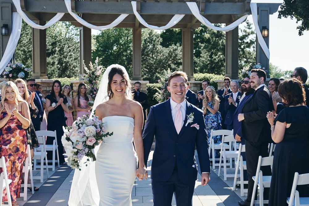 Bride and groom walk down the aisle at an outdoor wedding ceremony, guests applauding, bride holding a bouquet.