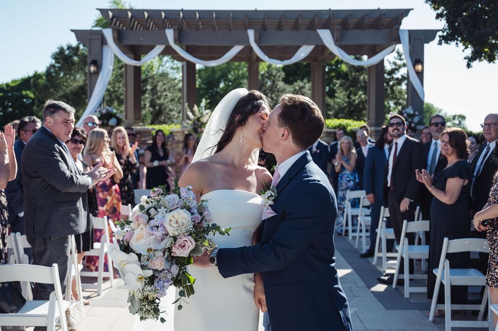 Bride and groom share first kiss at outdoor wedding ceremony with guests applauding and floral arch backdrop.