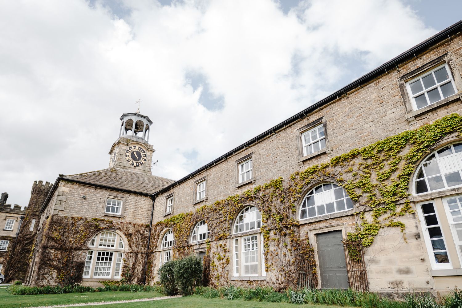 Historic stone building with ivy-covered walls, arched windows, and a clock tower under a cloudy sky.