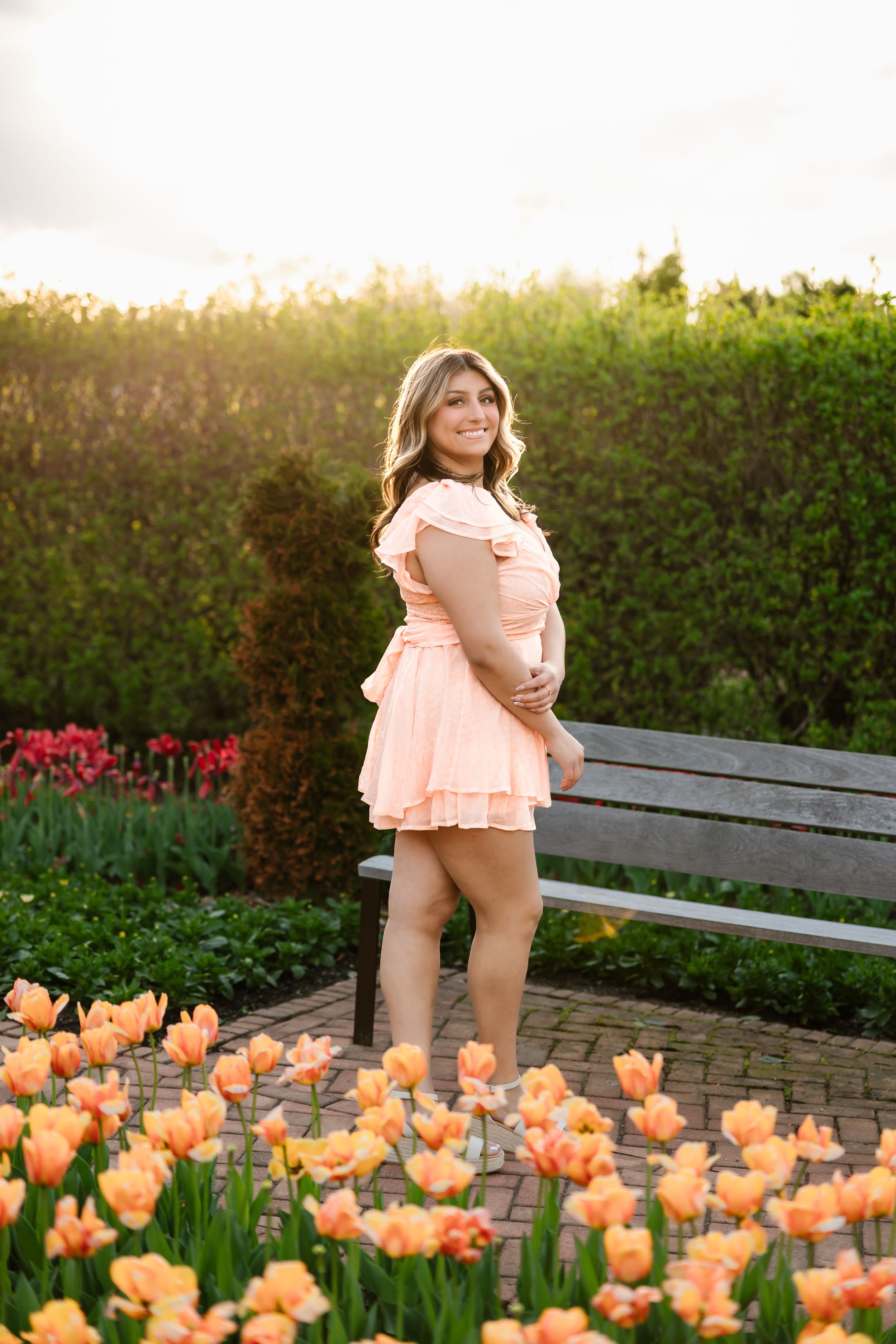 Young woman in peach mini dress sitting on bench surrounded by orange tulips in sunlit garden.