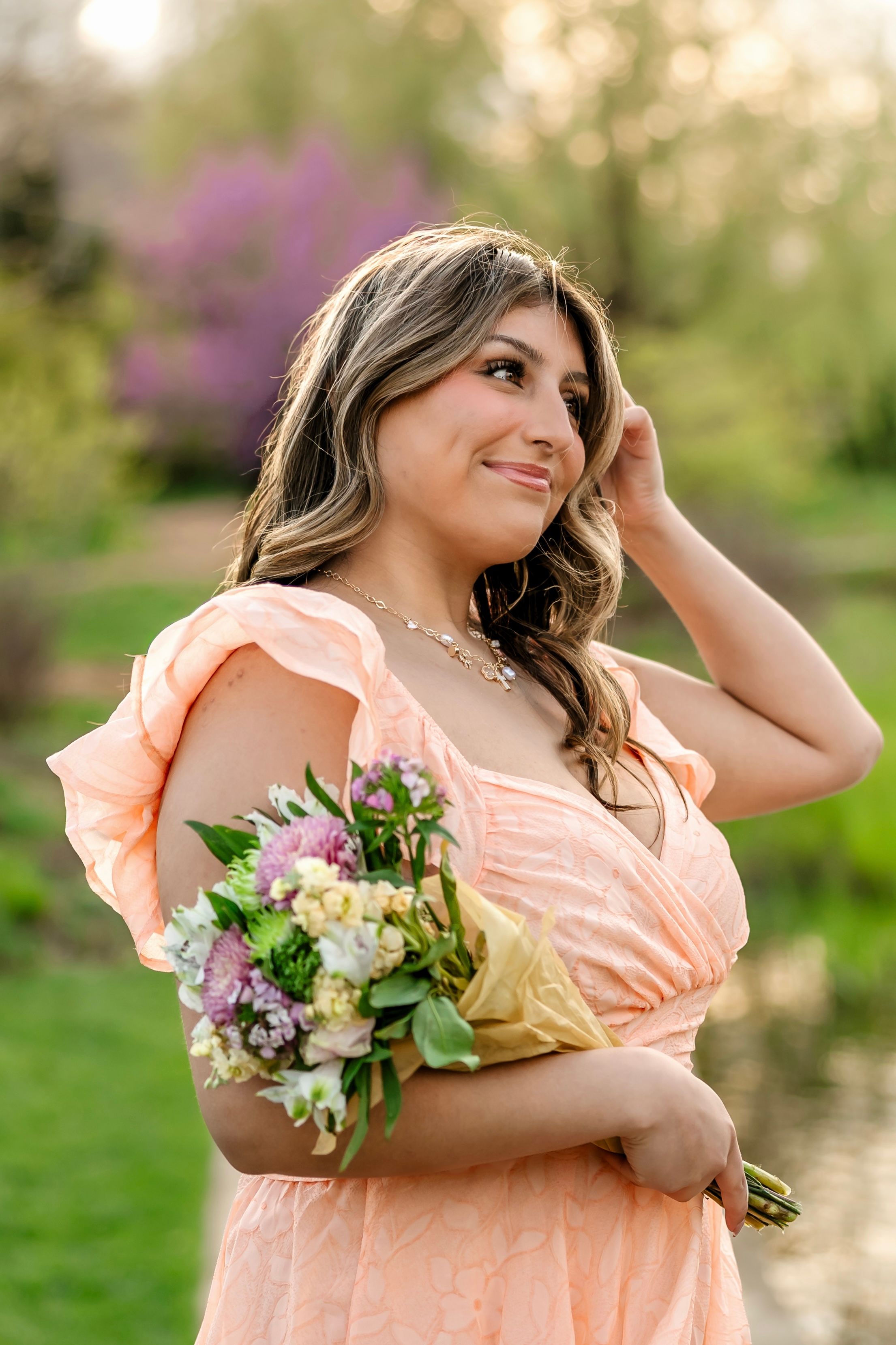 Young woman in peach off-shoulder dress holding spring bouquet in blooming garden at golden hour.