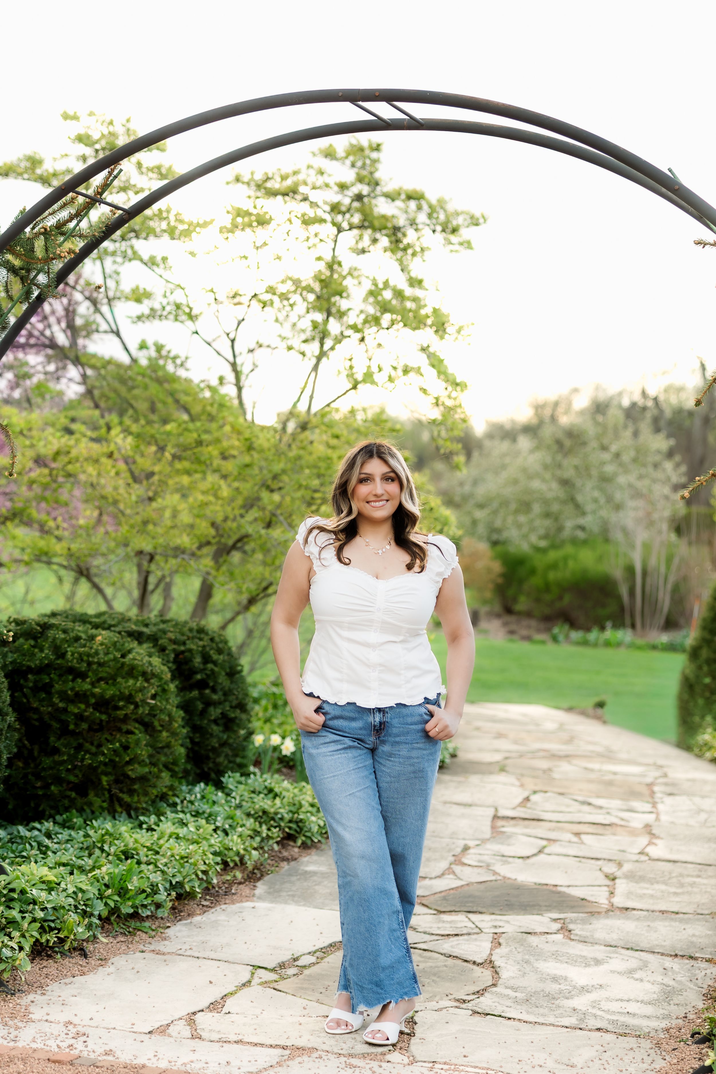 Smiling young woman in white top and jeans walking under metal arch on stone garden pathway.