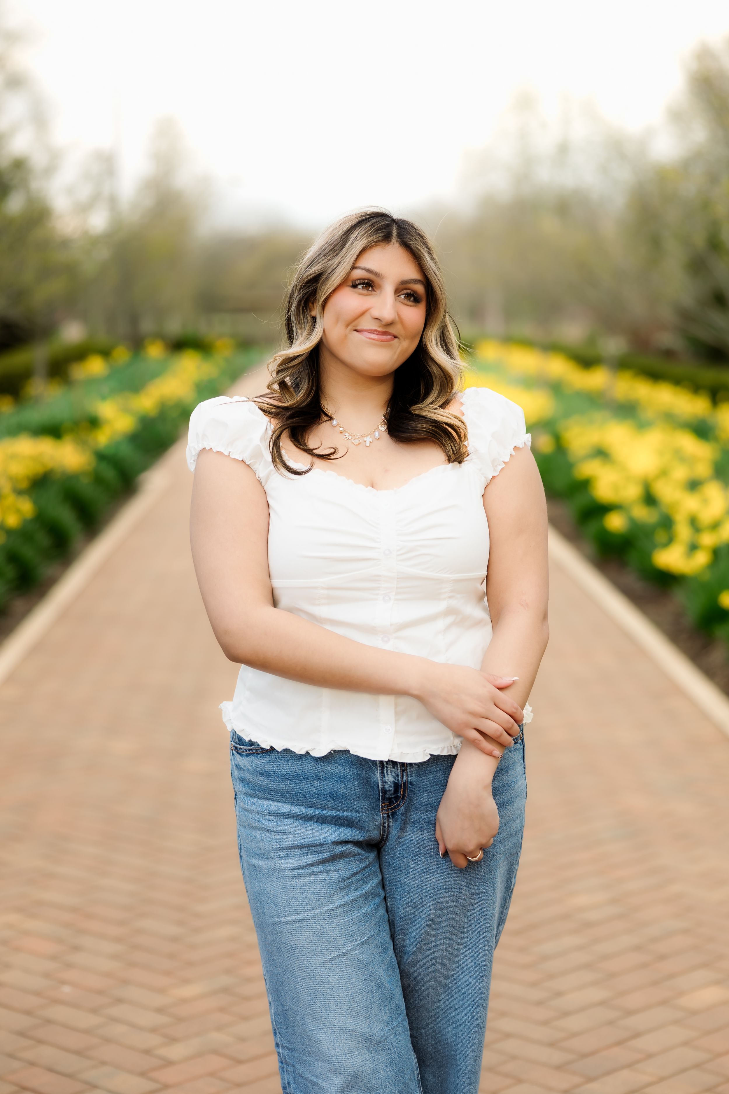 Smiling young woman in white top and jeans standing on brick path between rows of yellow tulips.