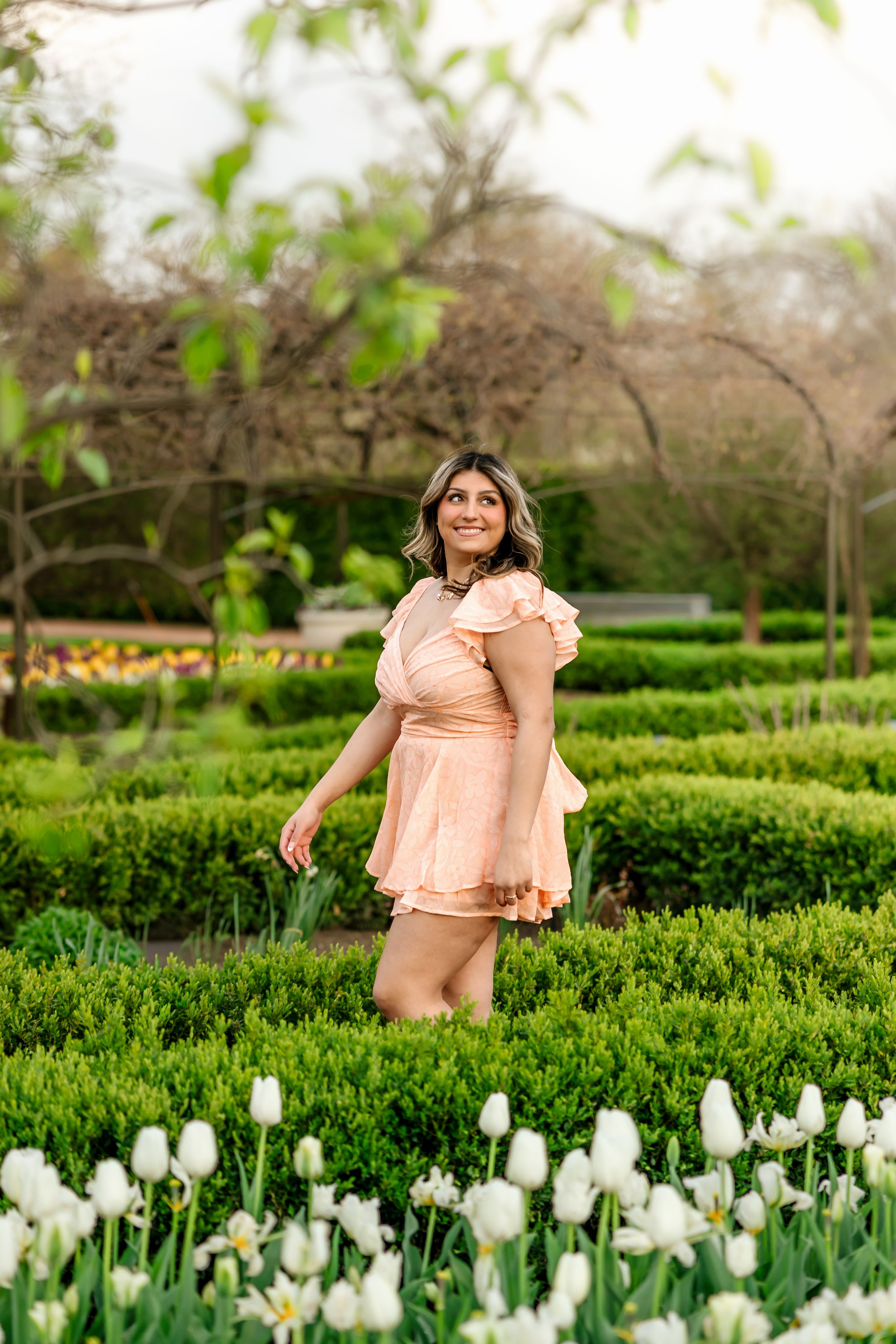 Young woman in peach mini dress posing among white tulips in a formal garden.