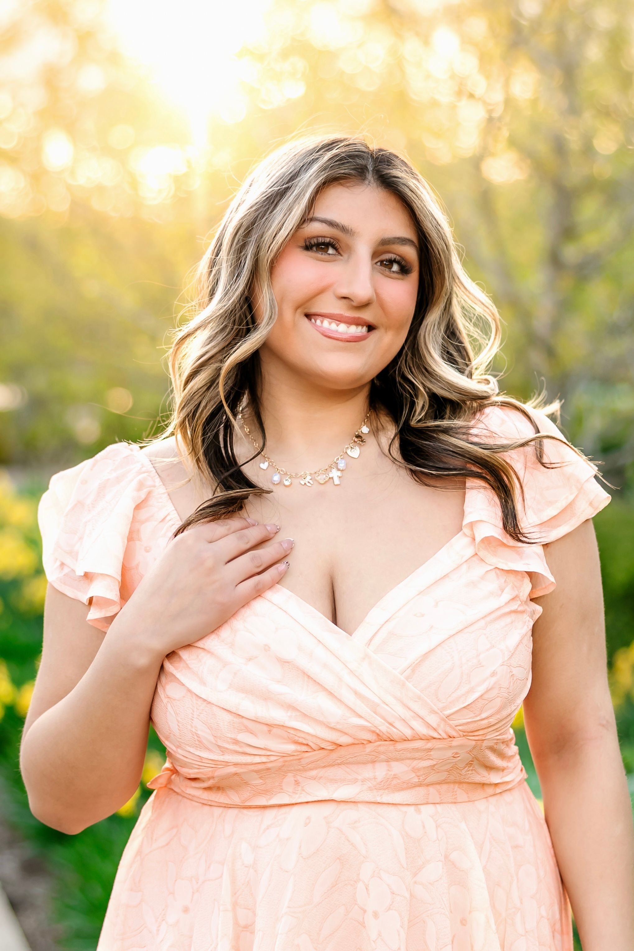 Young woman in peach ruffle dress with diamond necklace smiling outdoors in golden sunlight.