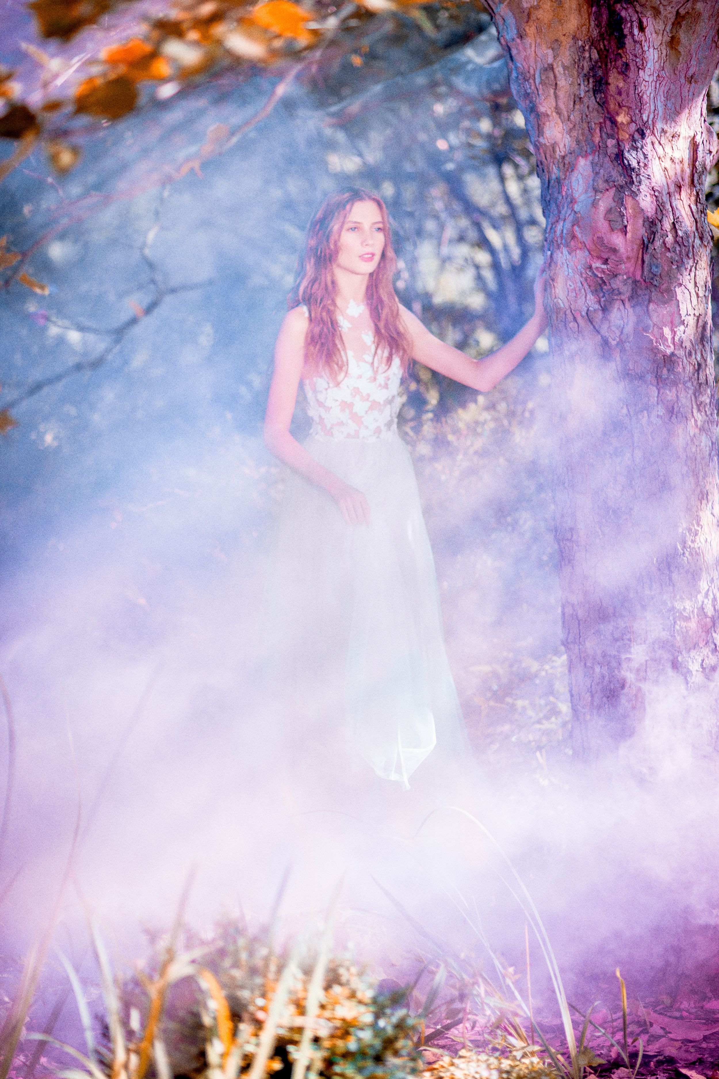 Woman in white floral gown standing by a tree surrounded by purple smoke in an enchanted forest setting.