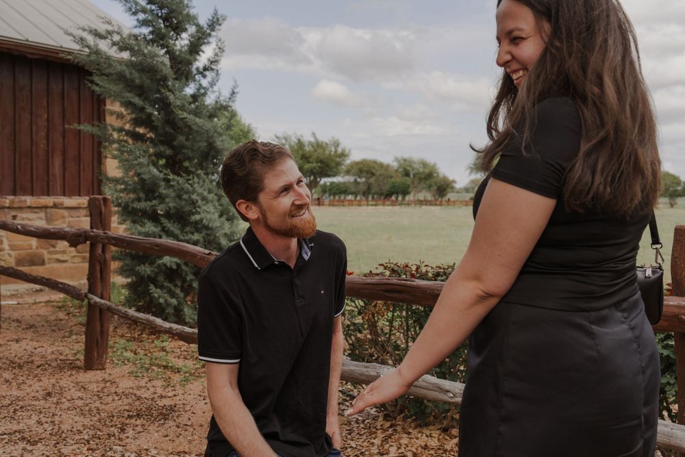 Man kneeling on one knee holding hands with a woman outdoors at a rustic farm venue during a proposal.