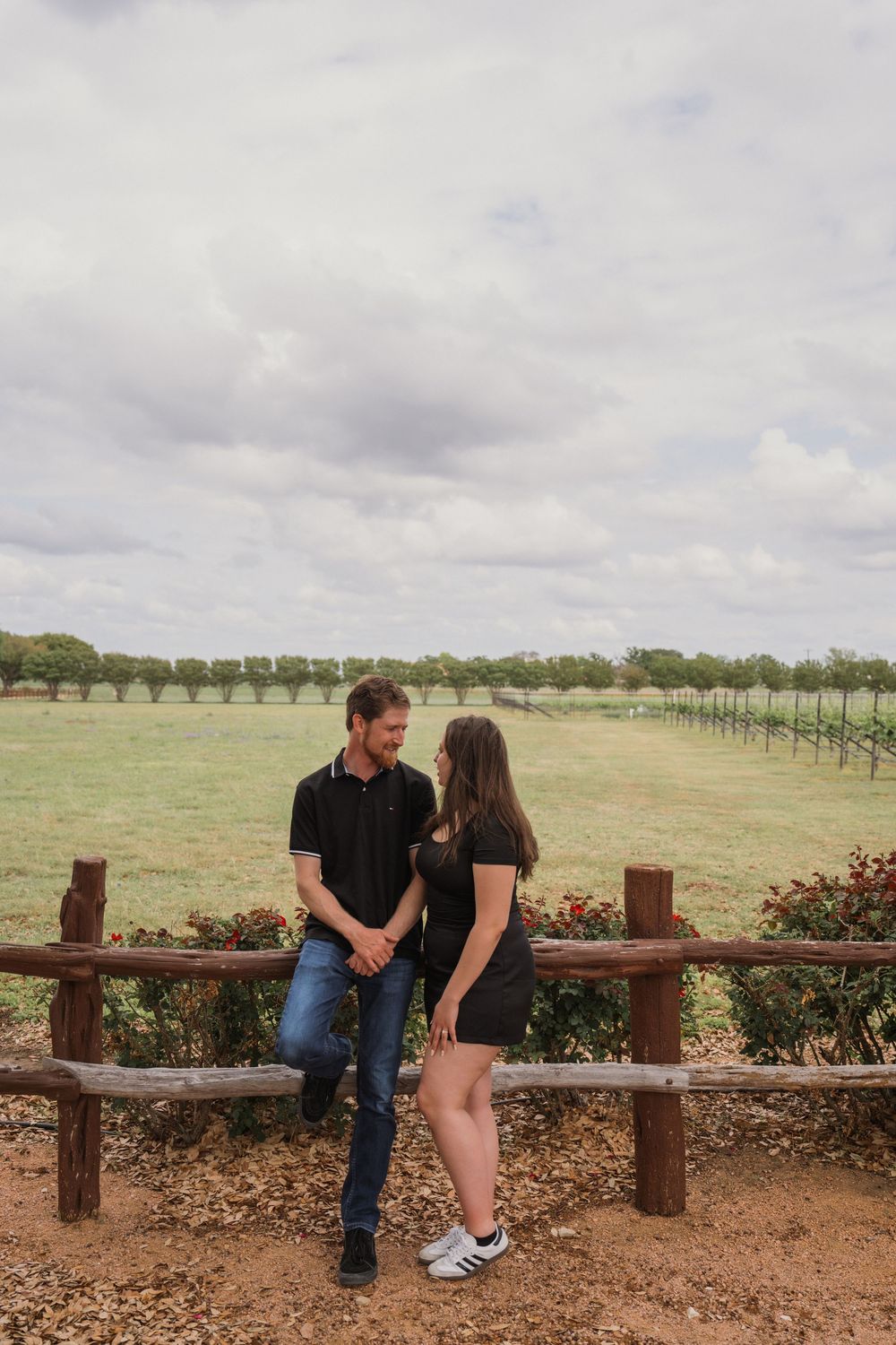 Couple in black outfits holding hands by rustic wooden fence with vineyard and cloudy sky backdrop.