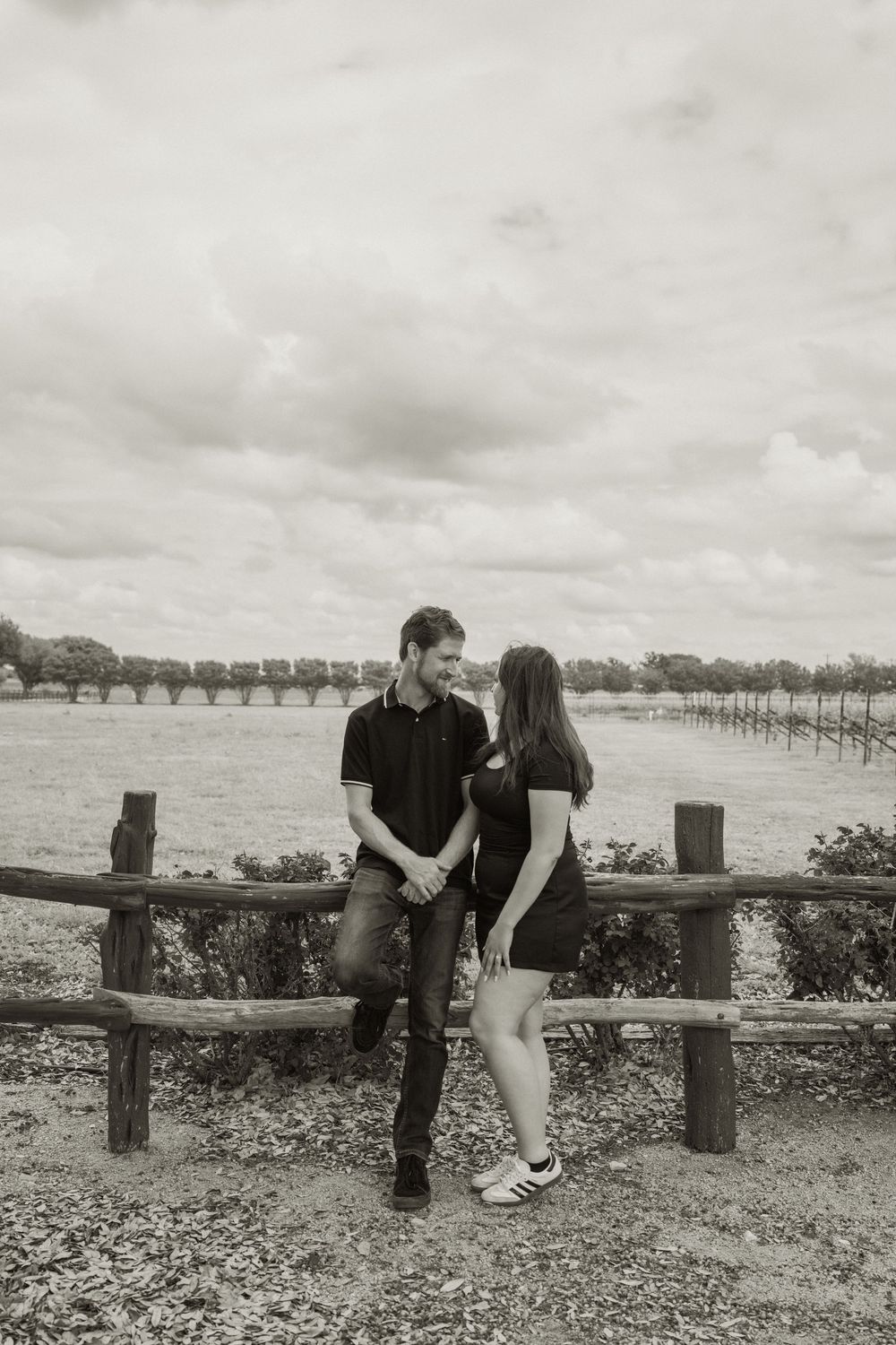 Black and white photo of a couple holding hands by a rustic wooden fence with vineyard rows in the background.