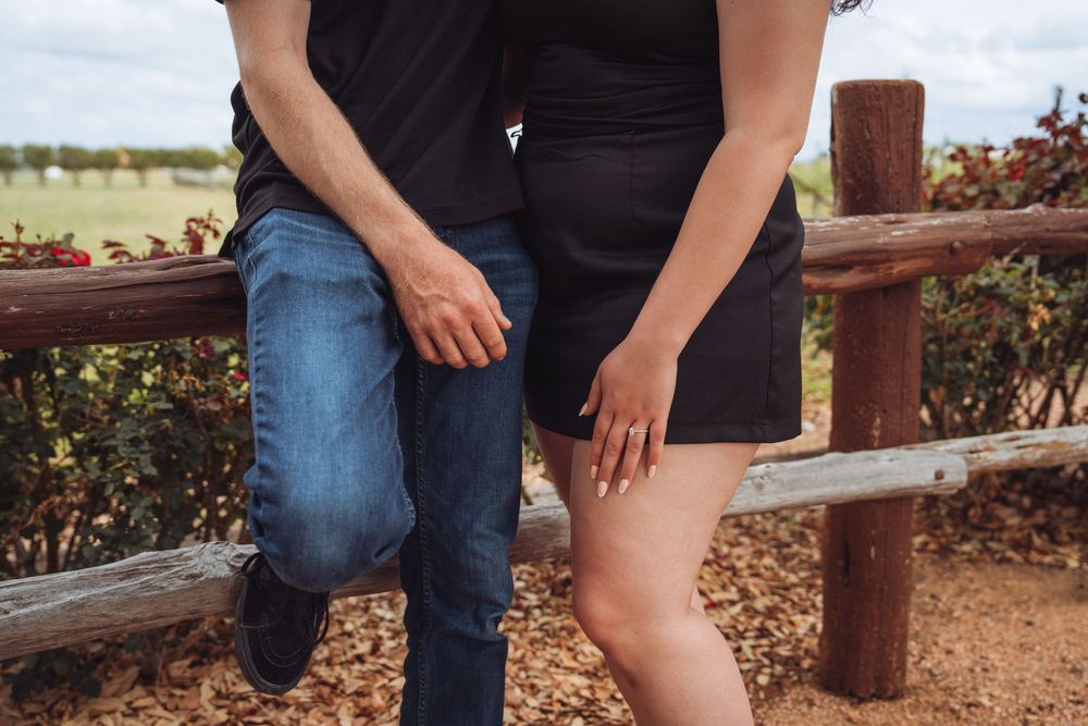 Couple standing close together by a rustic wooden fence outdoors, wearing casual black and denim outfits.