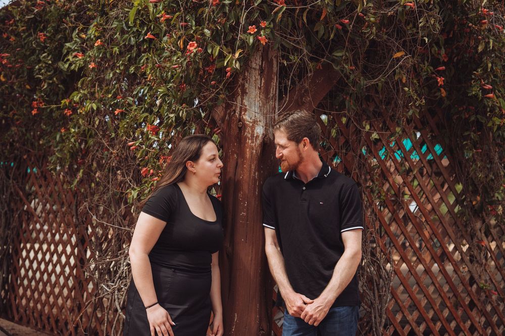 Couple in matching black outfits sharing a moment by a floral lattice wall.