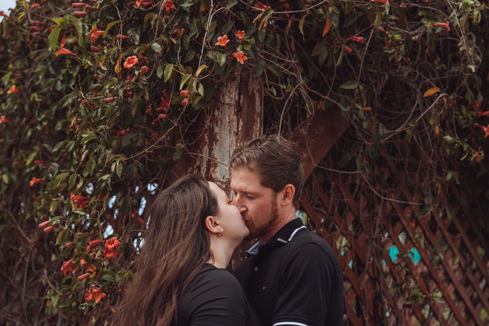 Romantic couple sharing a kiss under a flowering vine arch with red blooms in an outdoor garden.