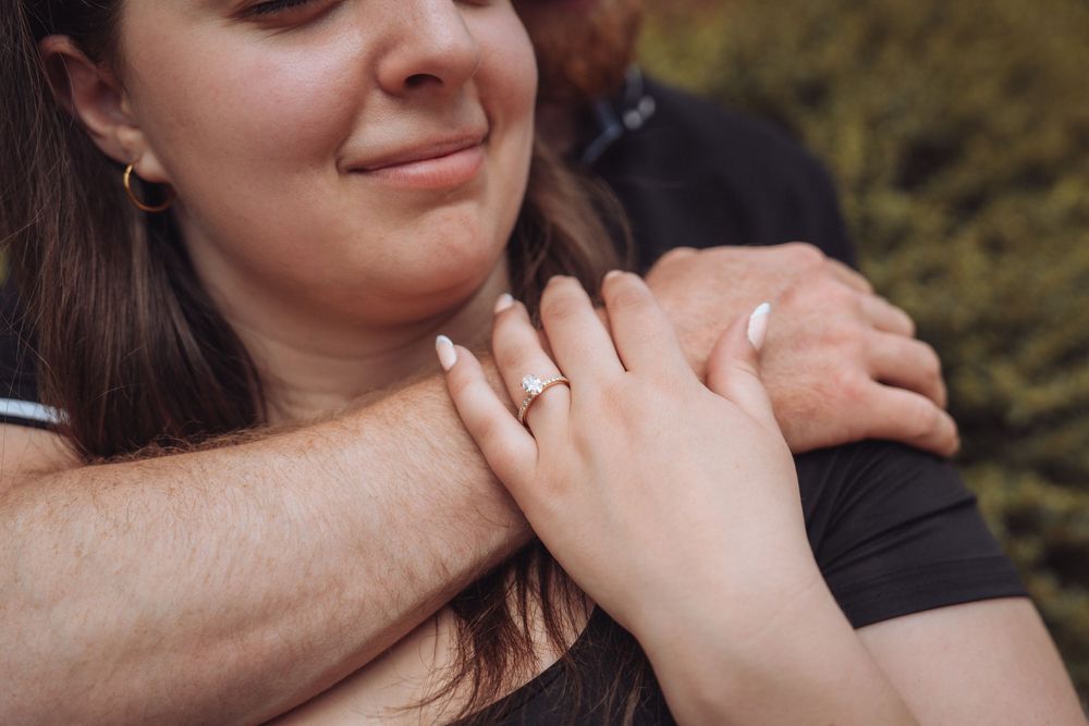 Close-up of a woman smiling while showing off her engagement ring as her partner embraces her from behind.