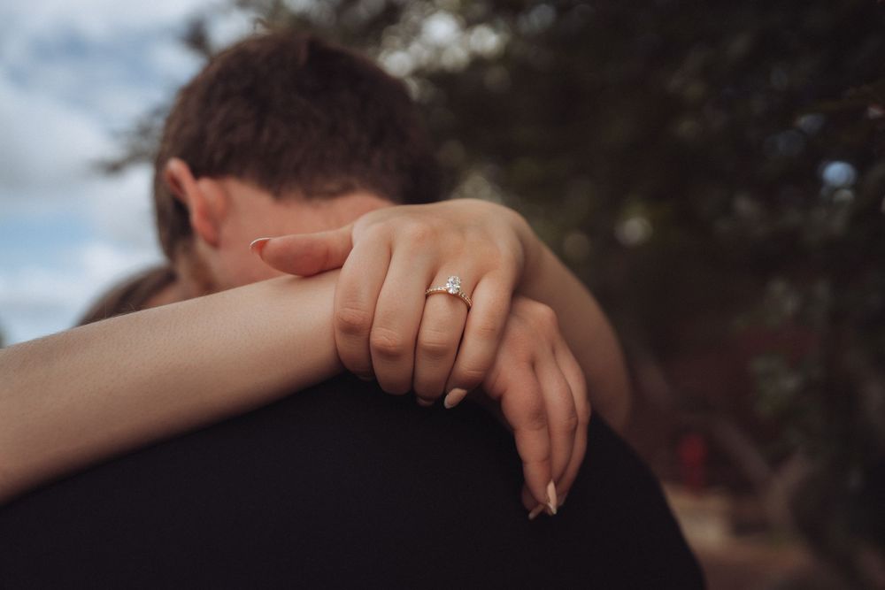 Close-up of a woman's hand with an engagement ring wrapped around her partner's shoulder in an embrace.