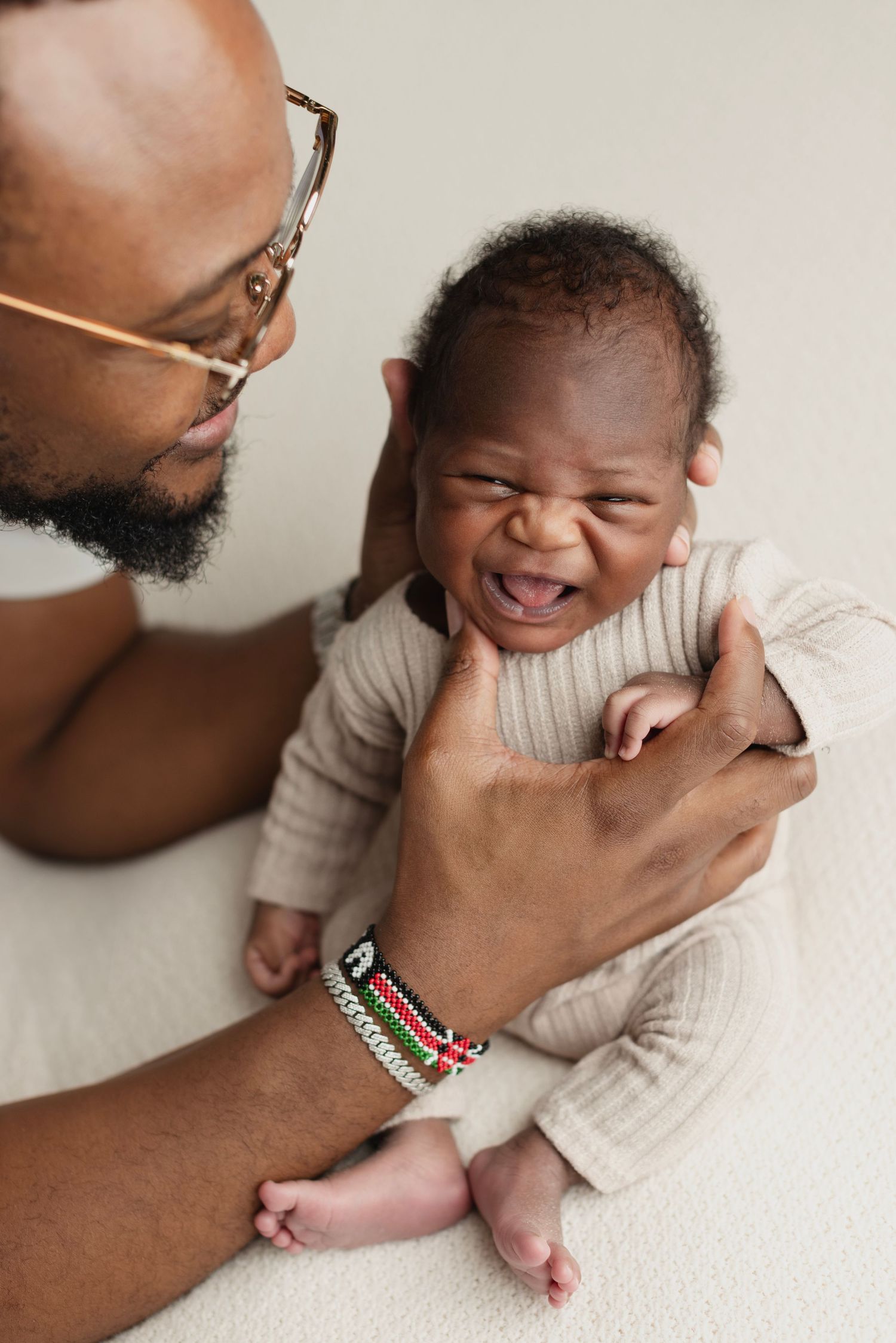A father with glasses and a beaded bracelet holds his crying newborn baby dressed in a beige ribbed outfit. Elizabeth Lang Photography Surre
