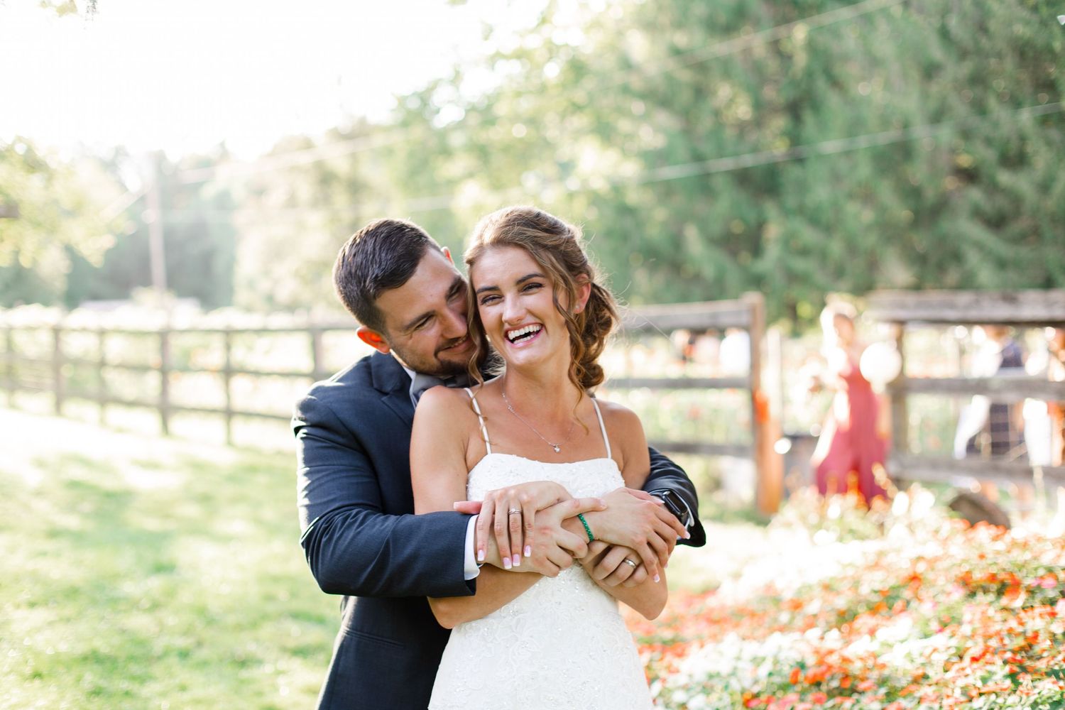 Happy bride and groom embrace outdoors at golden hour, with lush greenery and wooden fence backdrop.