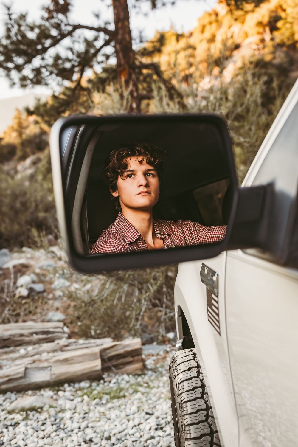 Young man with curly hair reflected in a white truck side mirror in a mountain setting in the inland empire for a senior photo session