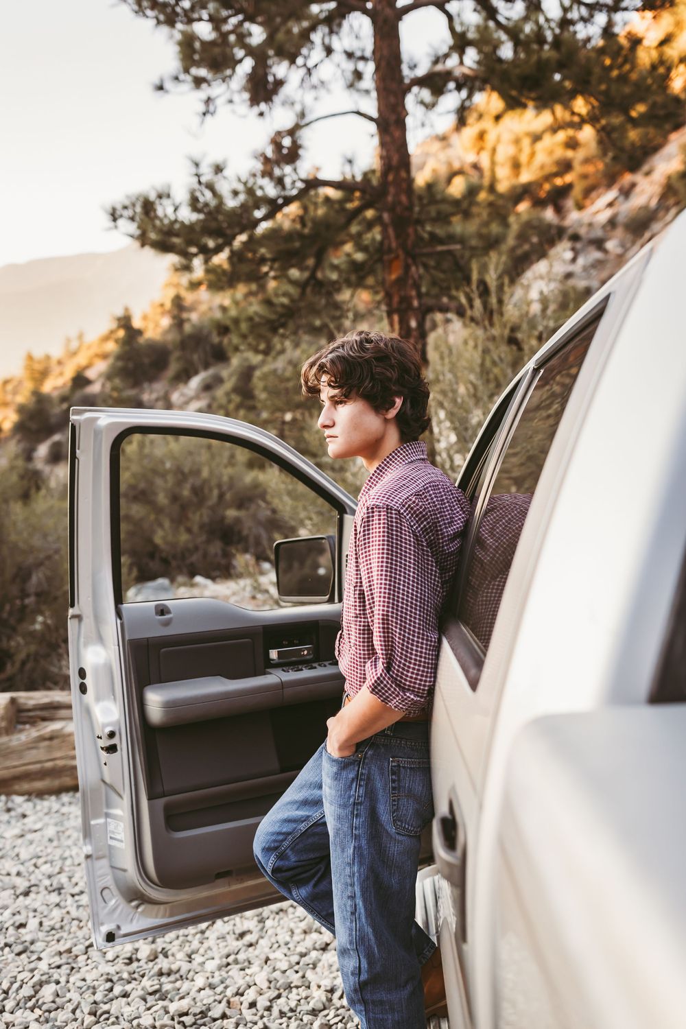 Young man in plaid shirt leaning against open white truck door in a rocky mountain landscape.