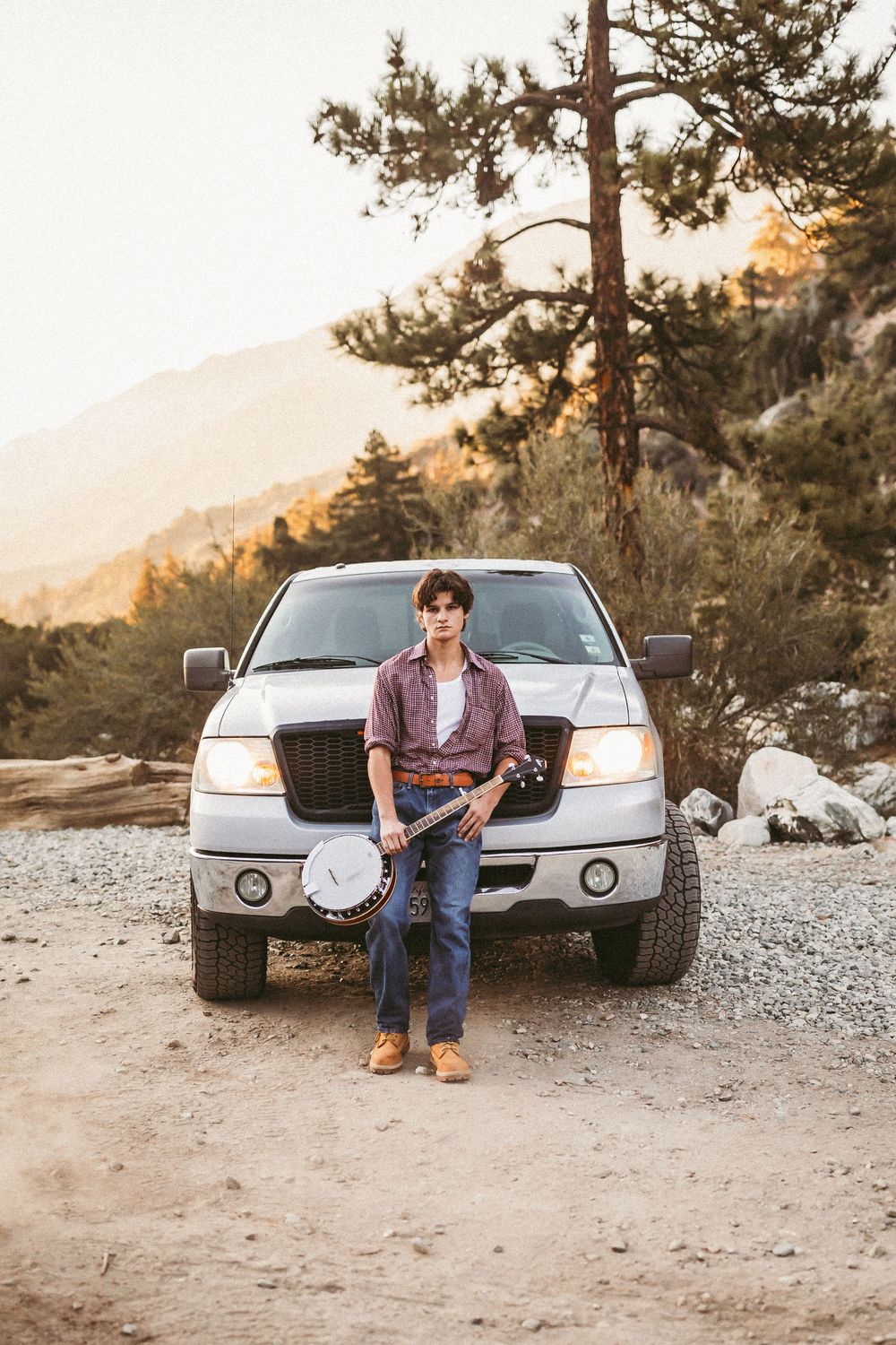 Young man holding a banjo leaning against a white pickup truck on a gravel road with mountains at sunset.