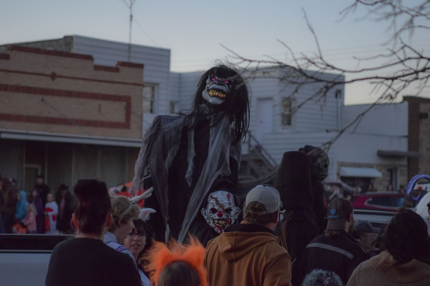 A person in a dramatic black reaper costume and mask towers over a Halloween parade crowd at dusk.