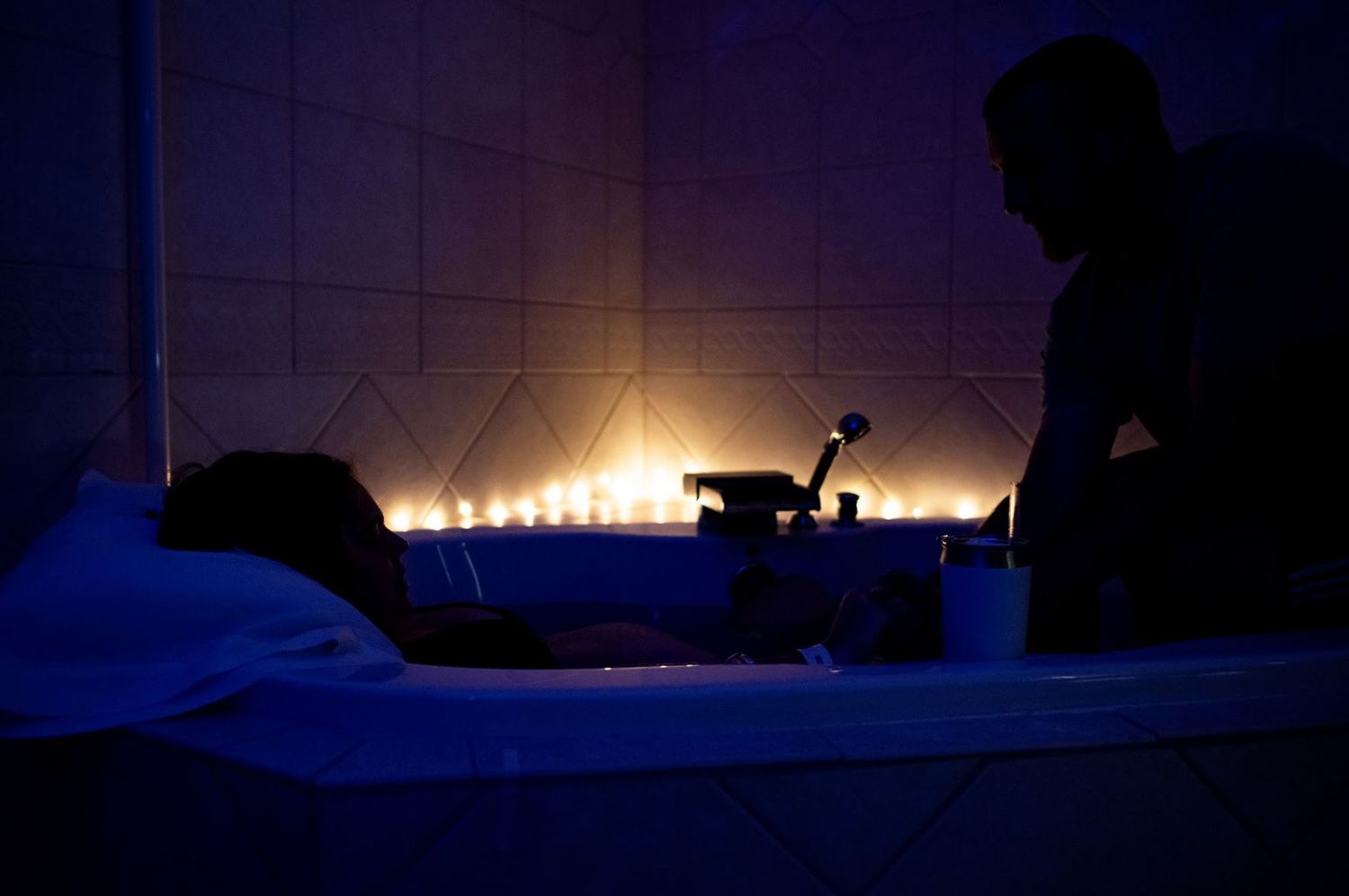 Silhouetted figure in dark room with glowing candlelight creating atmospheric mood against tiled wall.