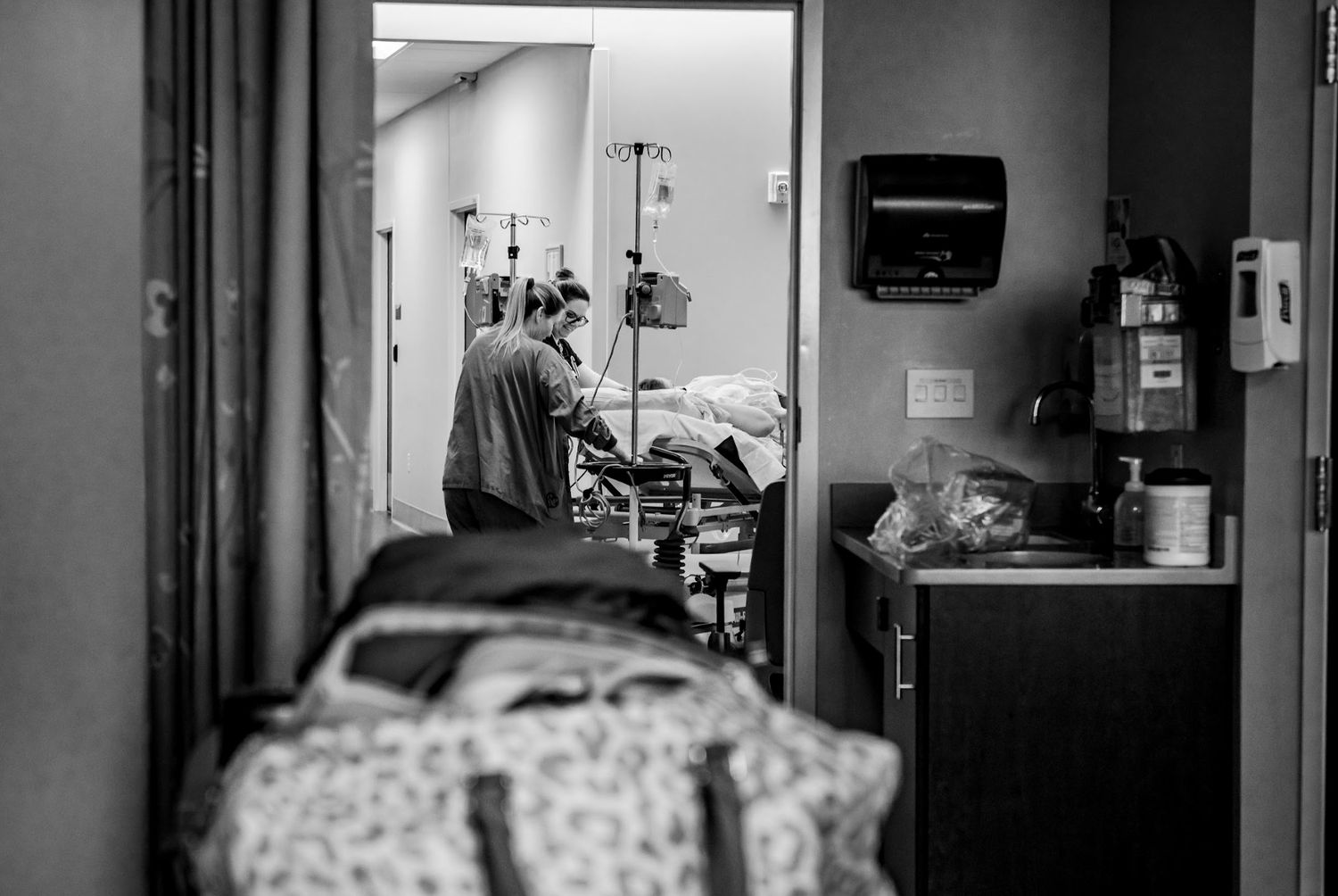 A black and white hospital room view through doorway shows medical equipment and staff.