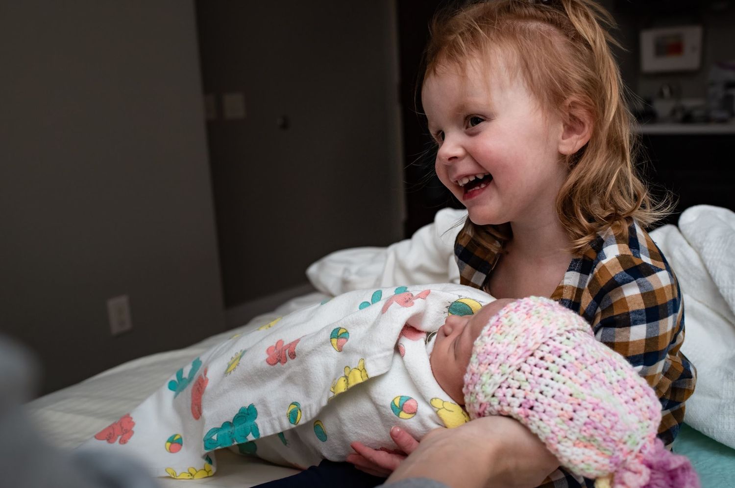 A young child smiles joyfully while holding her baby sister wrapped in floral blankets.