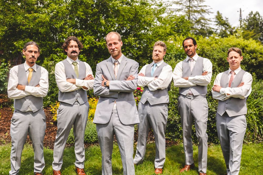 Six groomsmen in matching gray suits with colorful ties pose with arms crossed in a garden wedding photo.