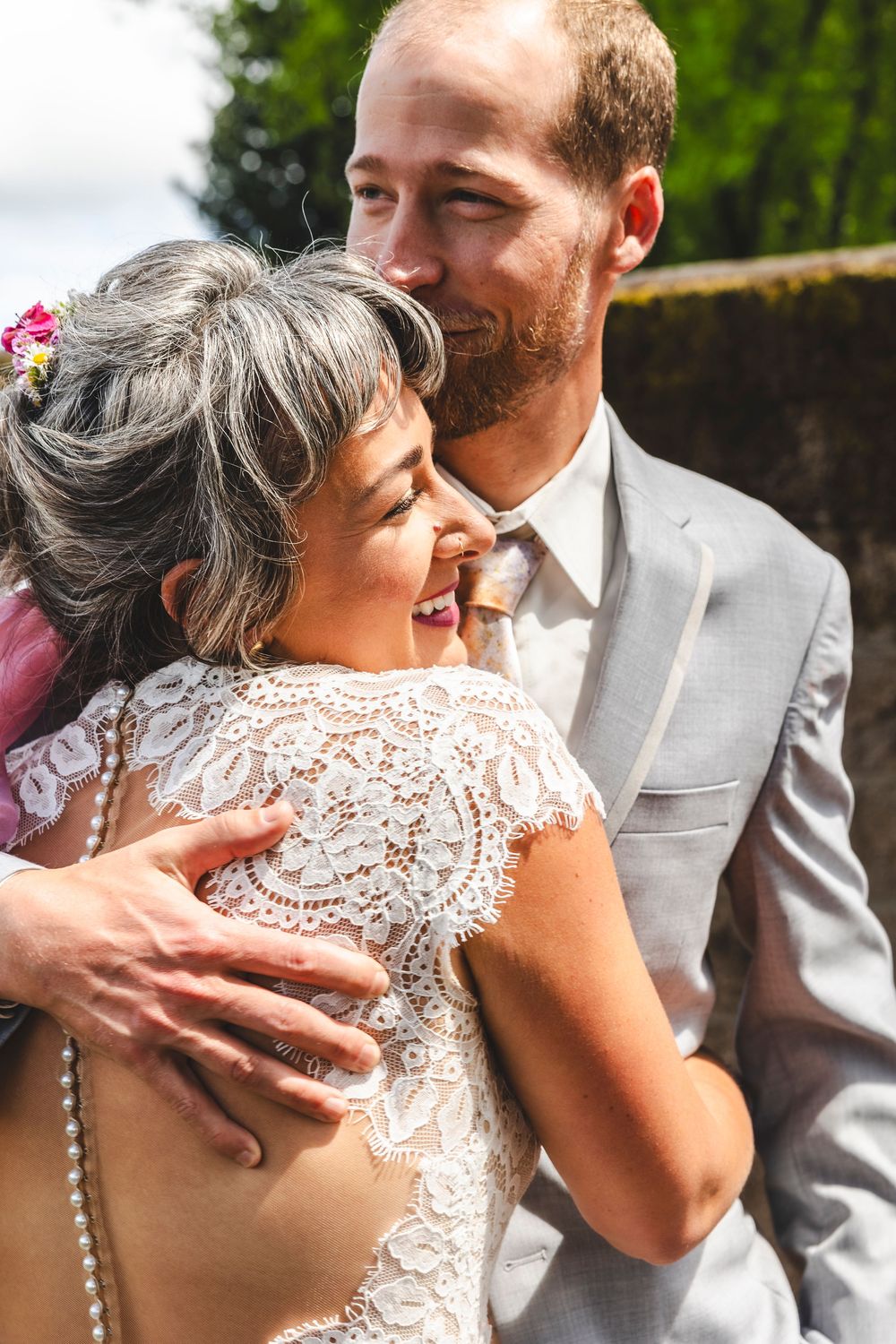A smiling bride in lace gown embraced by groom in gray suit at outdoor wedding ceremony.