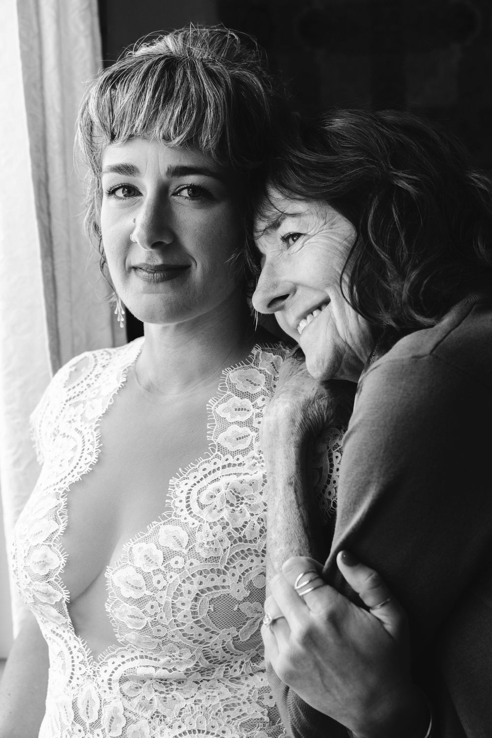 Black and white portrait of a bride in lace gown embracing an older woman near a window.