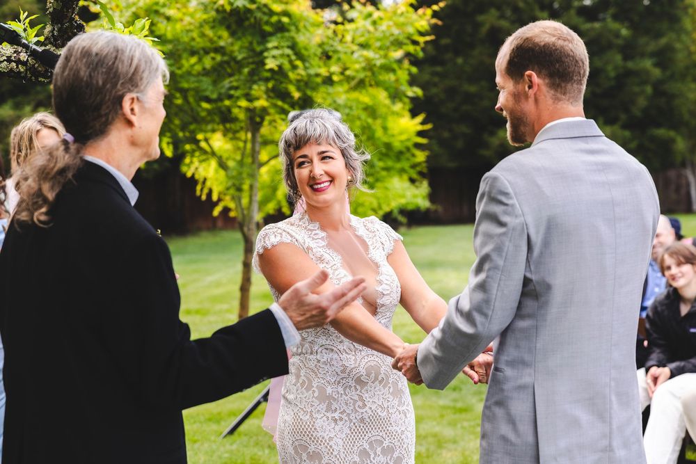 Bride in lace wedding dress laughing while holding hands with groom during outdoor wedding ceremony.