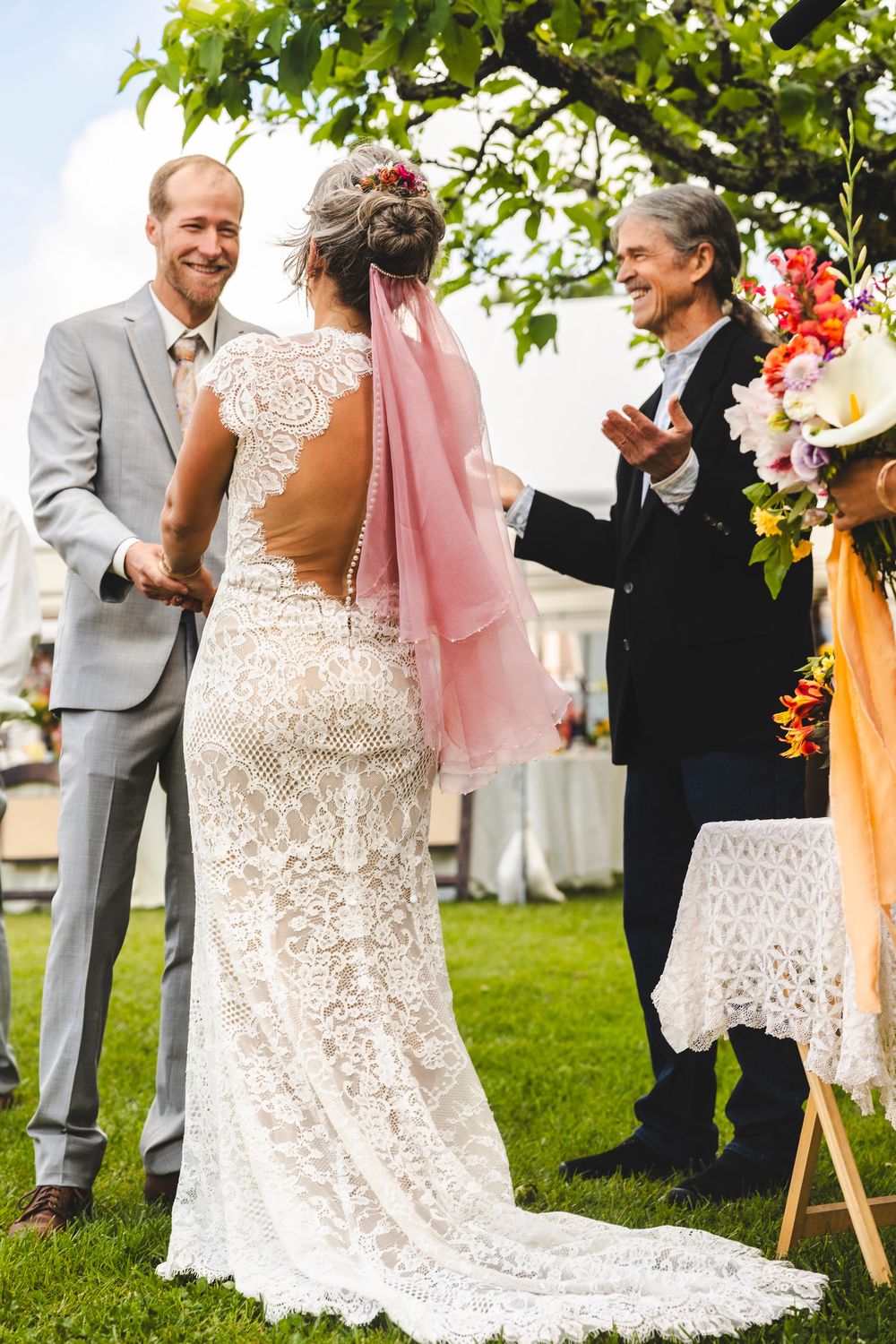 Bride in boho lace gown with pink veil holds hands with groom during outdoor garden wedding ceremony.