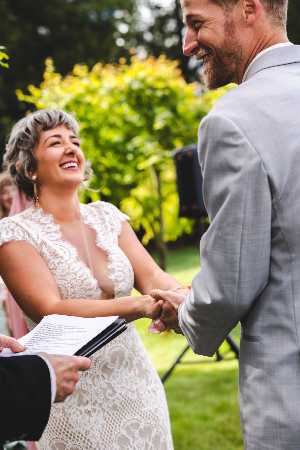 Smiling bride in lace dress exchanges vows while holding hands with groom at outdoor wedding.