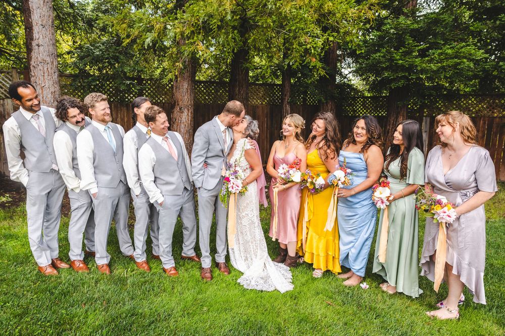 Wedding party poses together on lawn with groomsmen in gray suits and bridesmaids in colorful mismatched dresses.