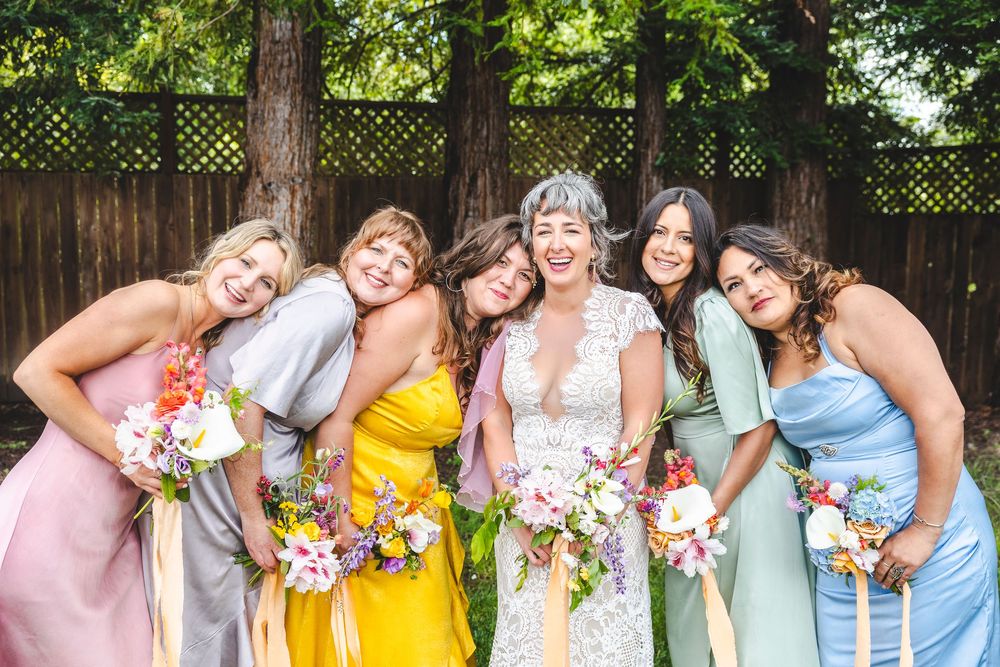 Bride surrounded by five bridesmaids in colorful mismatched dresses holding wildflower bouquets at outdoor wedding.