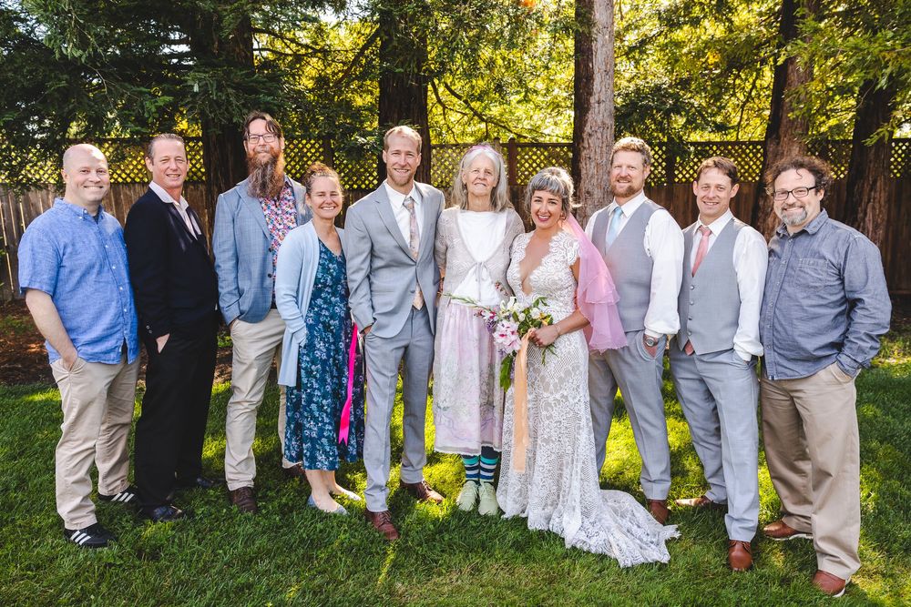 Wedding party group portrait with bride and groom surrounded by family and friends smiling outdoors in a garden.