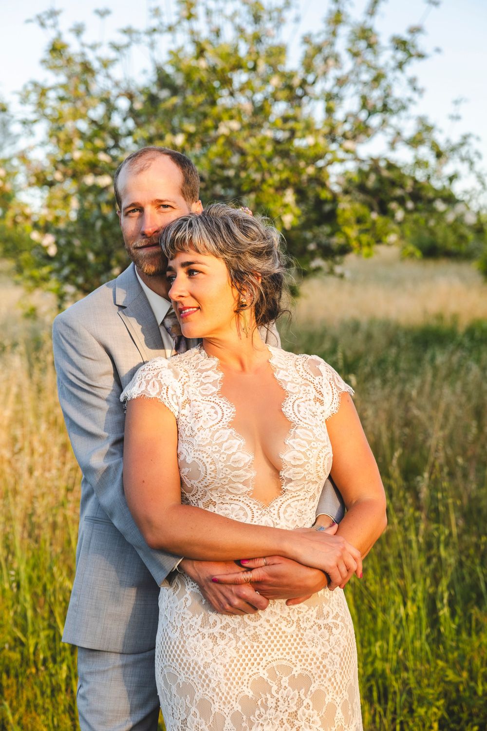 Bride and groom portrait in golden hour light, standing in a meadow with green trees behind.