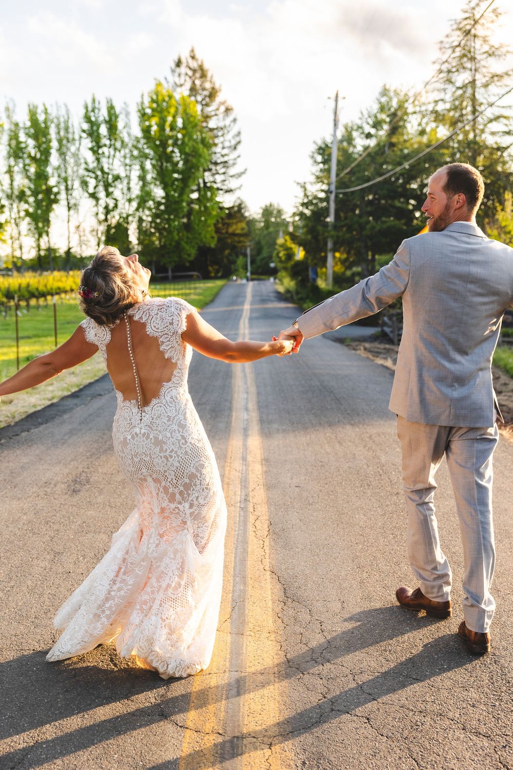 Bride and groom holding hands walking down a sunlit road, bride in lace backless gown, groom in gray suit.