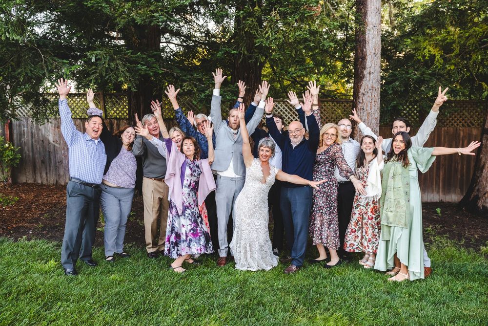 Joyful wedding group photo with guests raising arms in celebration in a backyard garden.
