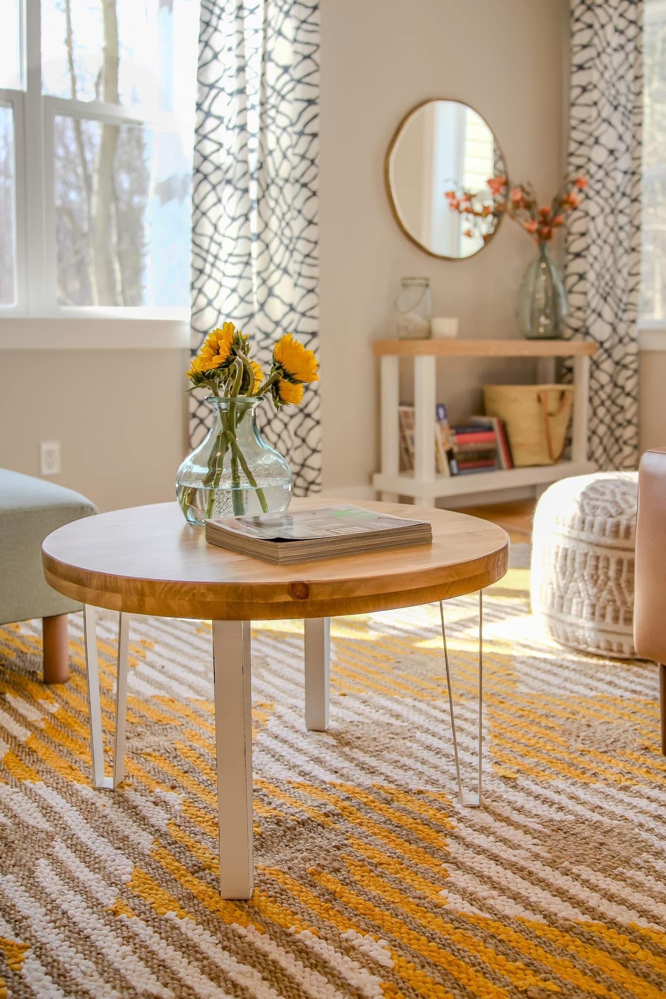 Bright living room with round wood coffee table, yellow flowers, patterned rug, and geometric curtains.