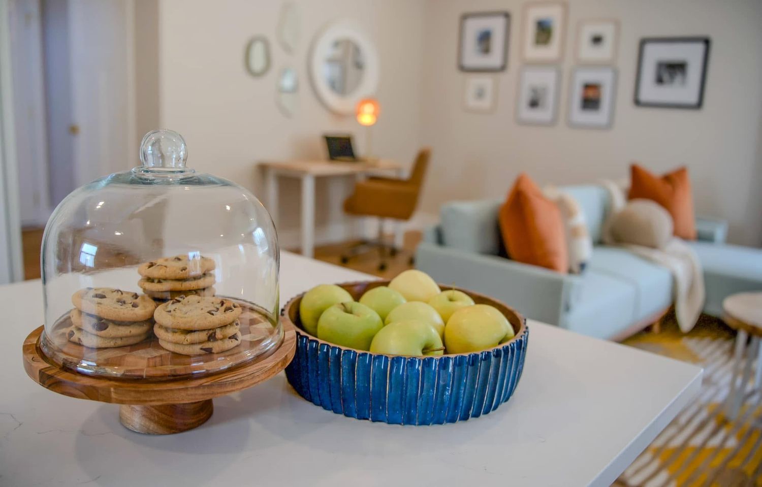 Modern living room with cookies under glass dome and green apples in blue bowl on white countertop.